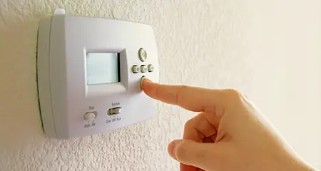 A person adjusting the temperature on a wall-mounted thermostat with their hand.