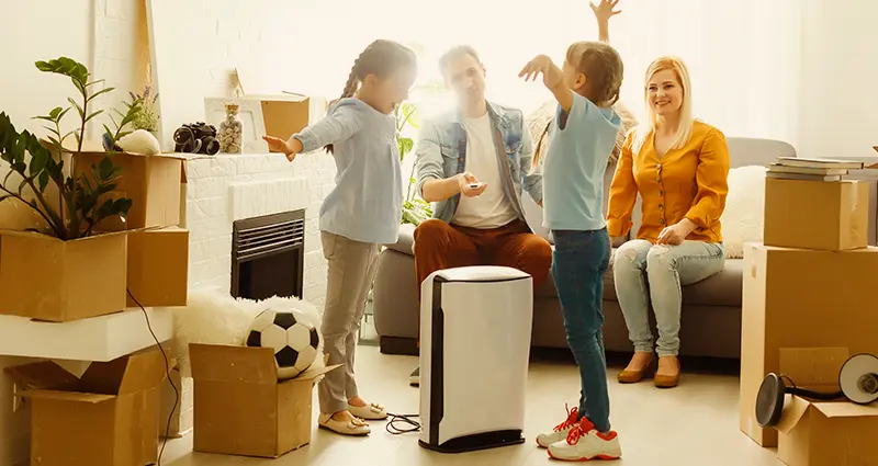 A family sits in a living room surrounded by moving boxes and a vacuum cleaner, indicating they are in the process of moving.