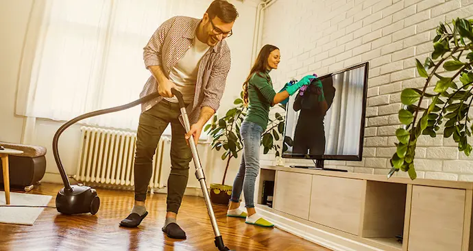 Couple cleaning living room. Husband is vacuuming floor and wife is dusting off the TV