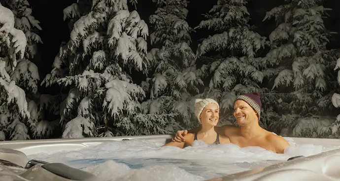 A couple relaxes in a hot tub surrounded by snow-covered trees, enjoying a serene winter landscape.