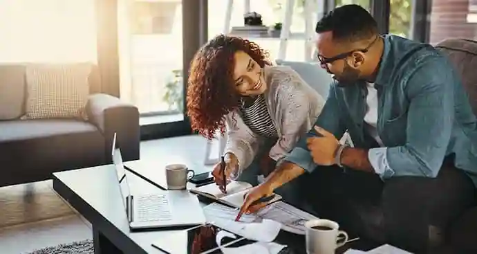 A man and woman are seated on a couch, both looking at each other with a laptop in front of them.