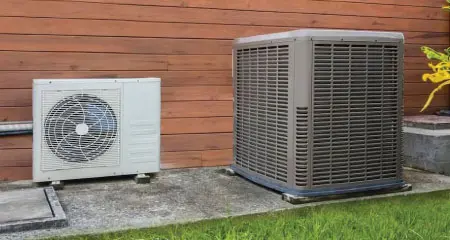 An outdoor heat pump and air conditioning unit, positioned on a concrete slab with greenery in the background.