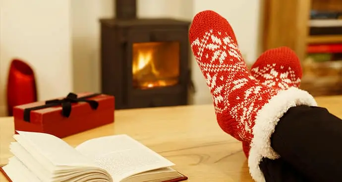 table with a giftbox next to an open book with feet on the table top resting, with a fireplace in the background