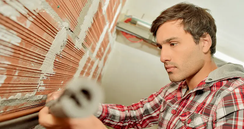 A man is repairing a wall while holding a pipe, focused on his task in a construction setting.