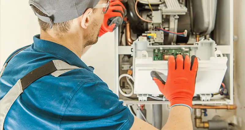 A man in a blue shirt and red gloves repairs a gas furnace, focused on his task in a well-lit workshop.
