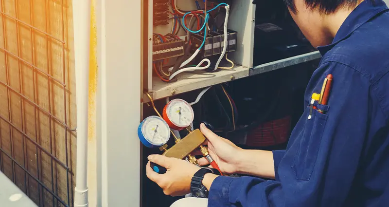 A man in a blue shirt repairs an air conditioner, focused on his work in a well-lit environment.