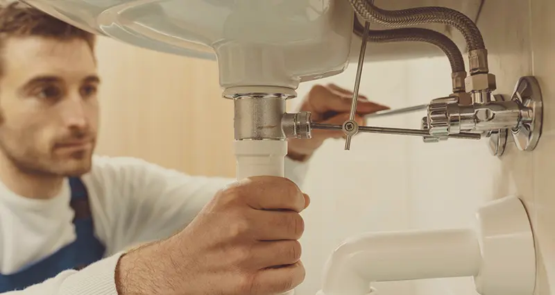 A man kneels beside a sink, repairing a faucet with tools in hand.