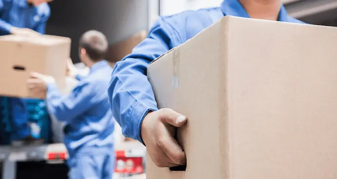 A man in blue overalls carries a large box, demonstrating strength and focus in his task.