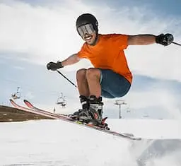 A man in an orange shirt and blue jean shorts skiing down a snowy slope, showcasing his skills by jumping through the air