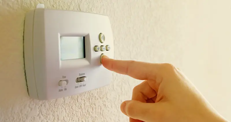 A person adjusting the temperature on a wall-mounted thermostat with their hand.