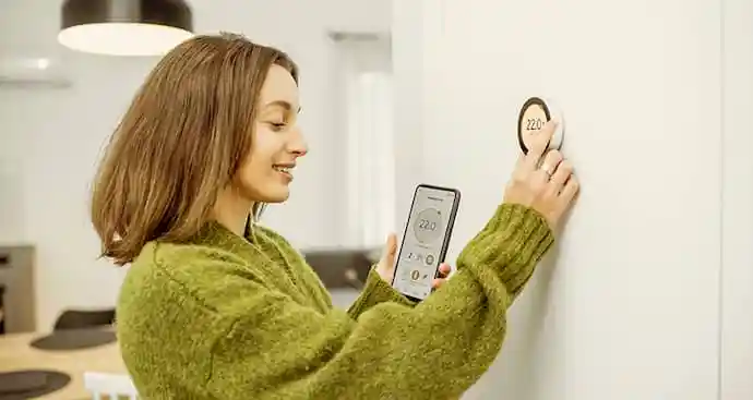  Woman adjusting smart thermostat while looking at mobile phone with climate control app open