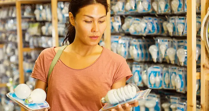 A woman in a hardward store, holding two different types of light bulbs figuring out which to buy