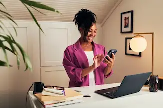 A woman multitasks at her desk, using her phone while working on her laptop.