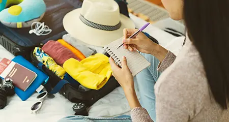 A woman sits on a bed with her luggage beside her, writing in a notebook.