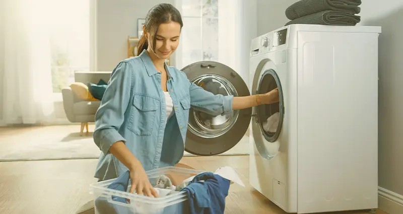 A woman is loading clothes into a front-loading washing machine in a bright laundry room.