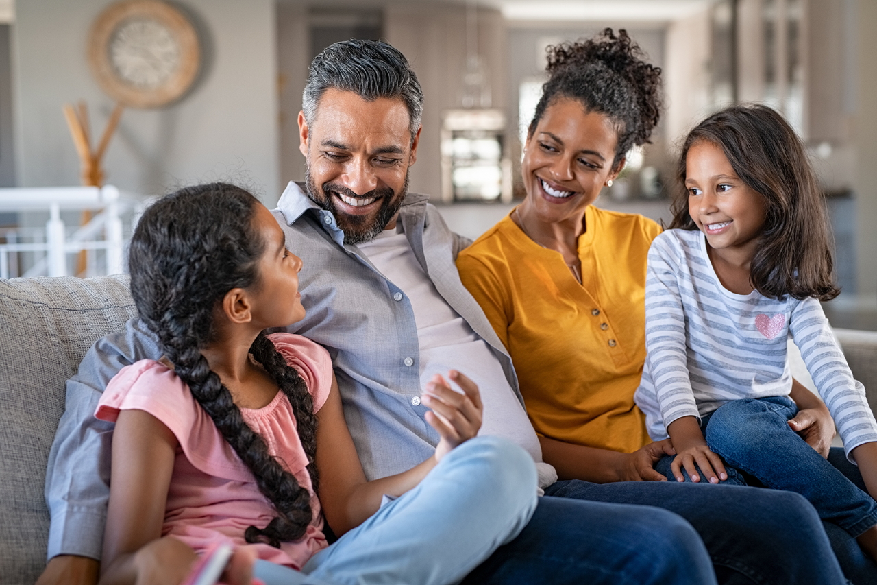 Cheerful family at home sitting on sofa listening to daughter. Happy couple with two girls relaxing at home together. Smiling indian parents talking to their daughter at home in the living room.