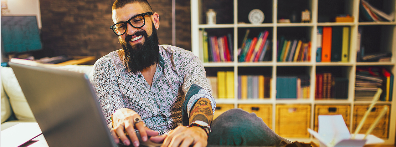 A man with glasses and a beard is seated at a desk, working on his laptop.