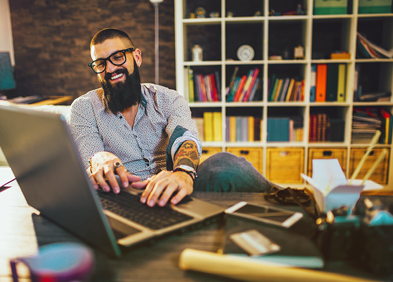 A man with glasses and a beard is seated at a desk, working on his laptop.