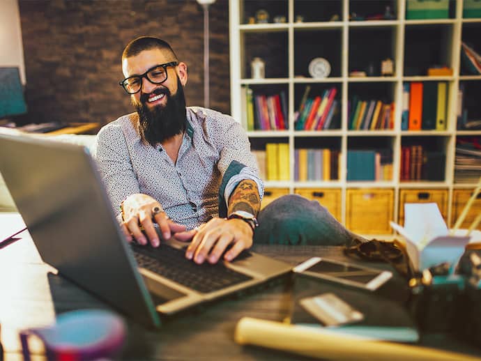 A man with glasses and a beard is seated at a desk, working on his laptop.