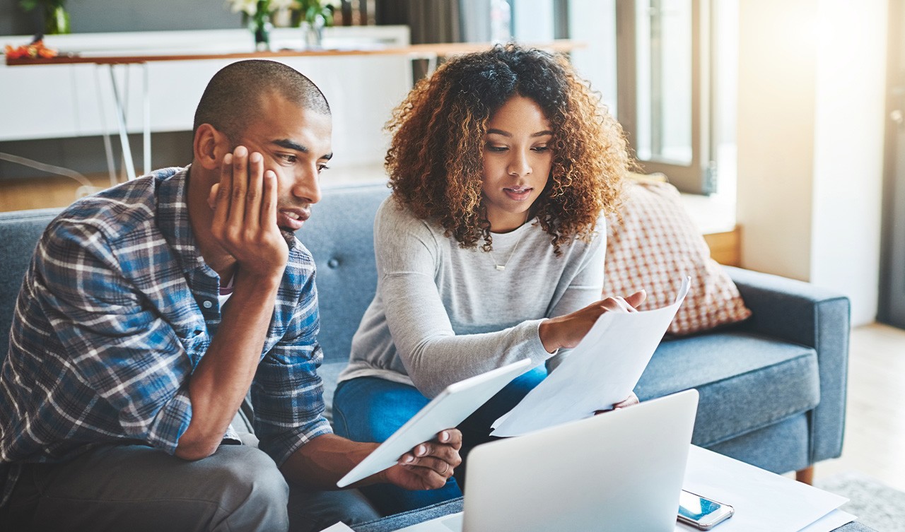 Two individuals collaborating on budgeting, surrounded by papers and a laptop in a comfortable living space.