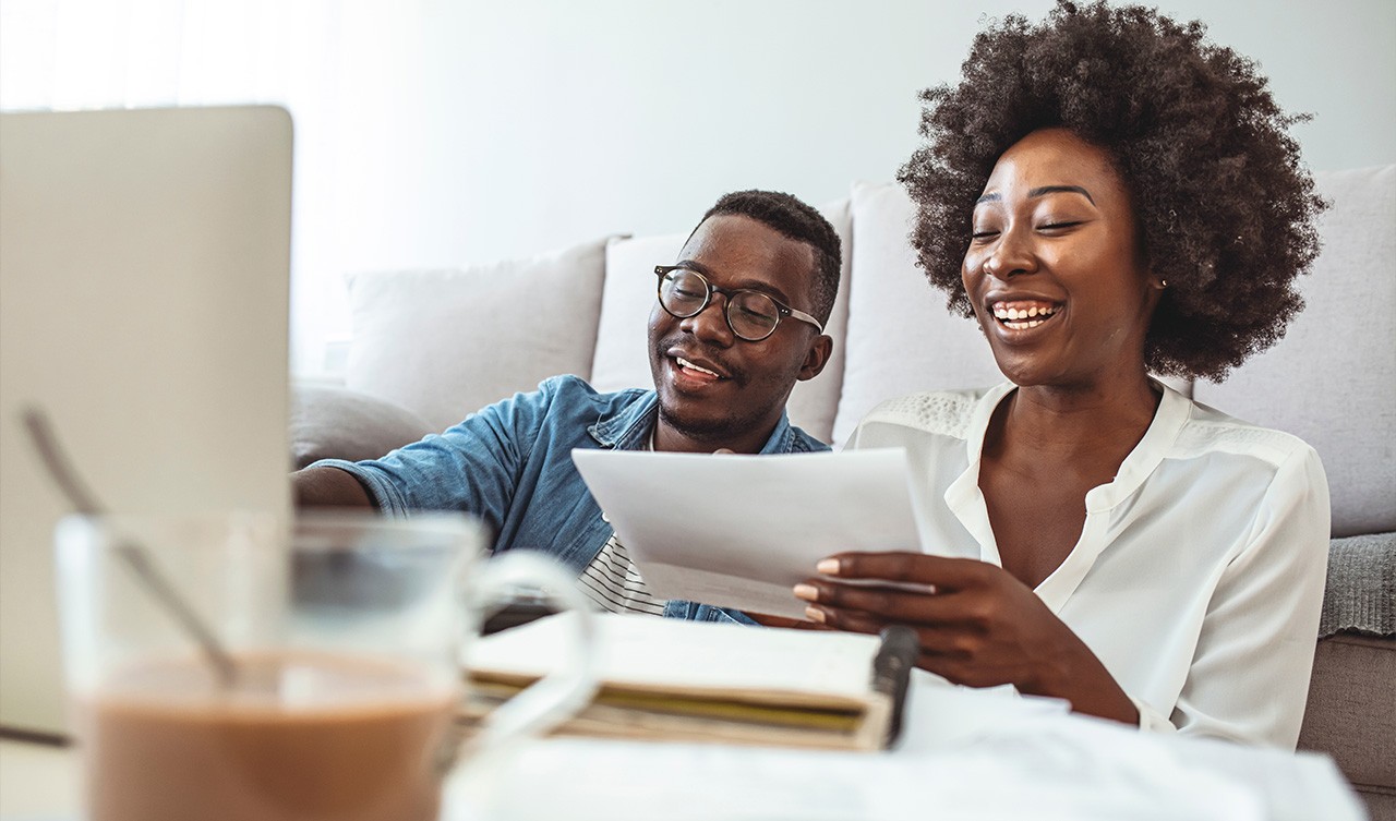 A young couple sits in a living room, focused on a laptop and reviewing a paper together.