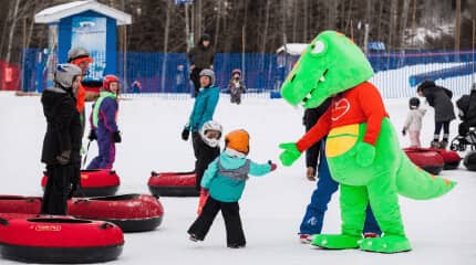 A cheerful dinosaur mascot stands beside people on a snowy slope, adding excitement to the winter landscape