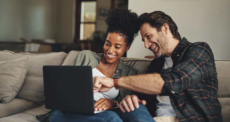 A man and woman sit together on a couch, focused on a laptop screen in front of them.