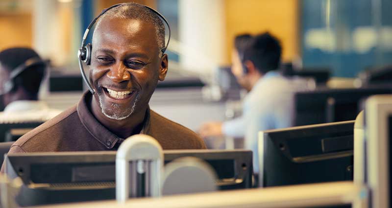 A smiling man wearing a headset, sitting at a desk in a call center, engaged in a conversation.