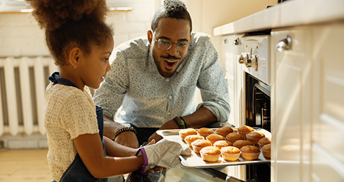 Young girl taking muffins out of the oven with her dad looking on.
