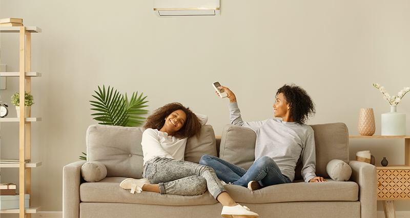 Mother and daughter sitting on couch with mom pointing remove at portable air conditioner