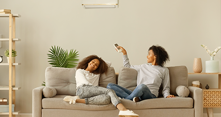 Mother and daughter sitting on couch with mom pointing remove at portable air conditioner
