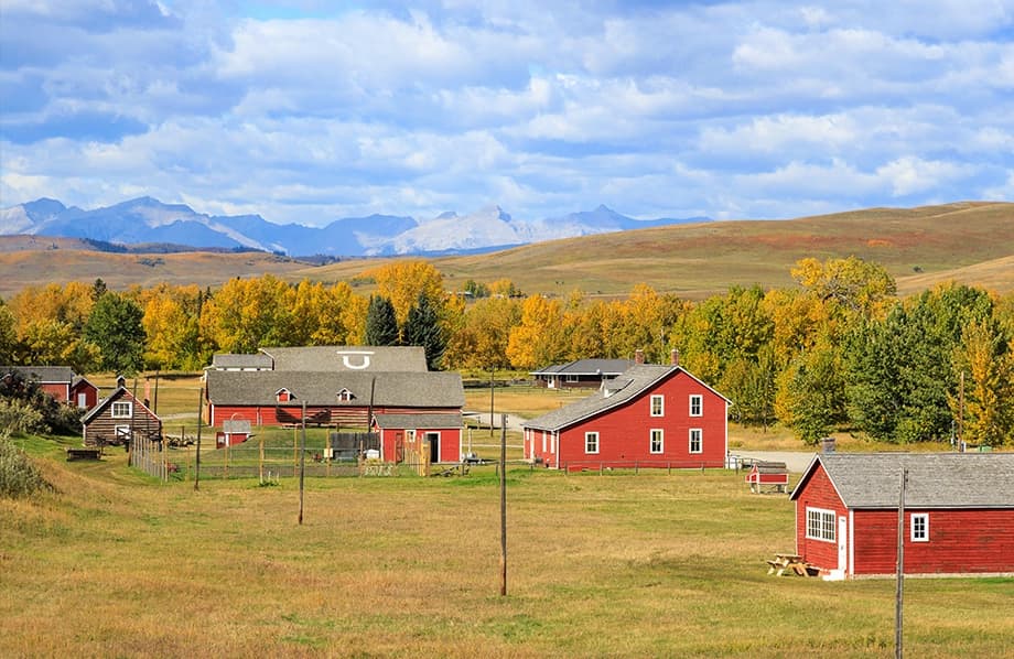 Red barns and houses stand on a grassy field with autumn trees and distant mountains under a partly cloudy sky.