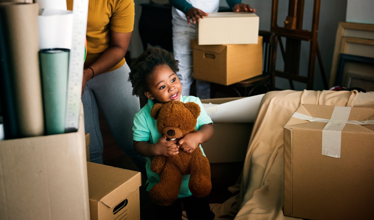 little girl with teddy bear exploring her new house with parents in the background unpacking boxes