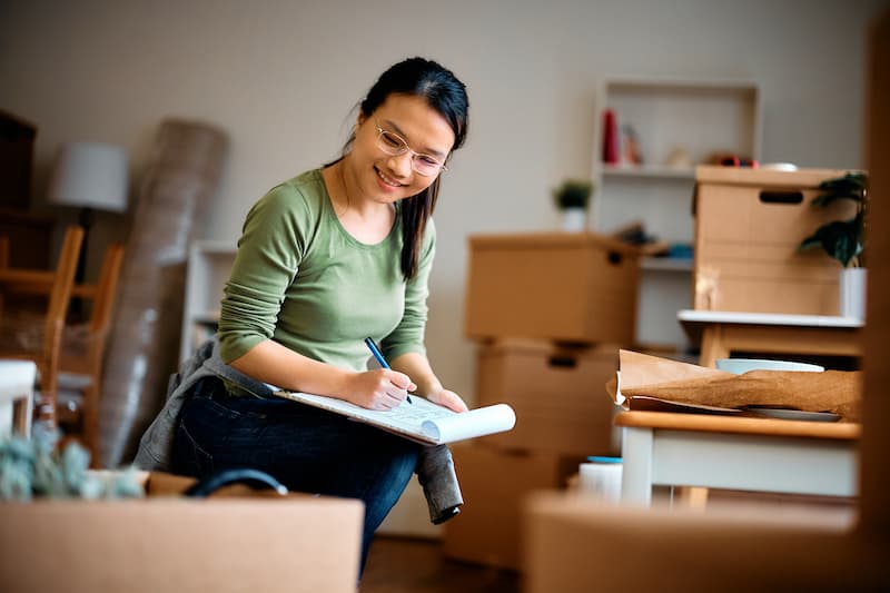 A woman sits in a cluttered room surrounded by boxes and papers, appearing focused on her task.