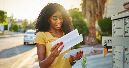 A woman stands next to a mailbox, holding a piece of paper, appearing to read or review its contents