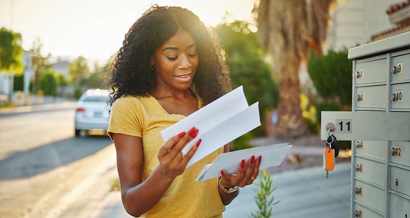 A woman stands next to a mailbox, holding a piece of paper, appearing to read or review its contents