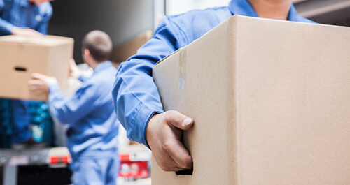 A man in blue overalls lifts a box, highlighting his effort and concentration as he moves forward