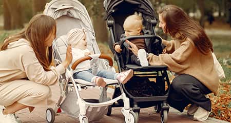 Two women are smiling at each other near a stroller with a baby, set against a backdrop of trees and sunlight.