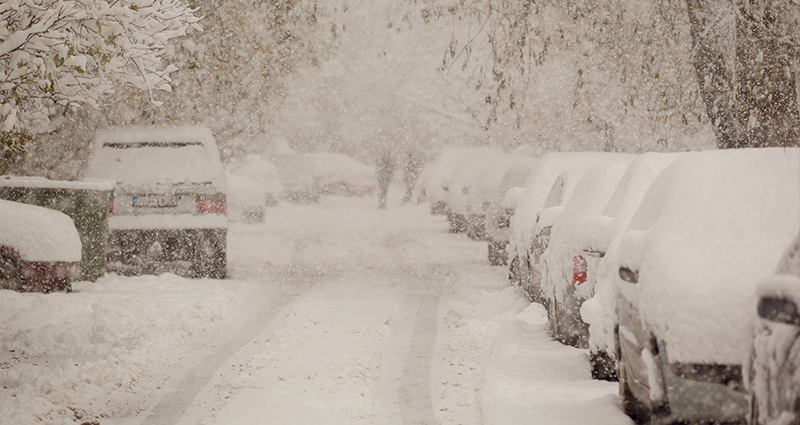 A snowy street with parked cars, their surfaces blanketed in white snow, creating a serene winter scene.