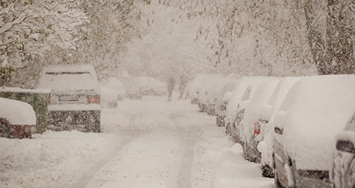 A snowy street with parked cars, their surfaces blanketed in white snow, creating a serene winter scene.