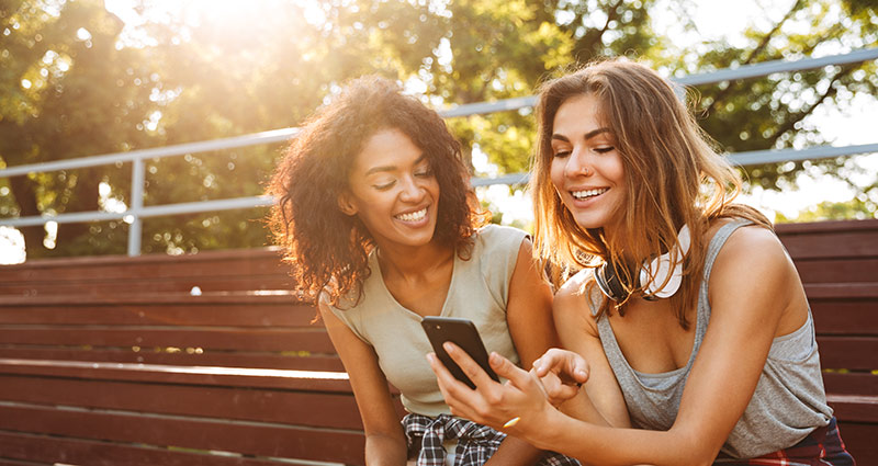 Two young women sit on a bench, focused on their cell phones, sharing a moment of connection in a public space.