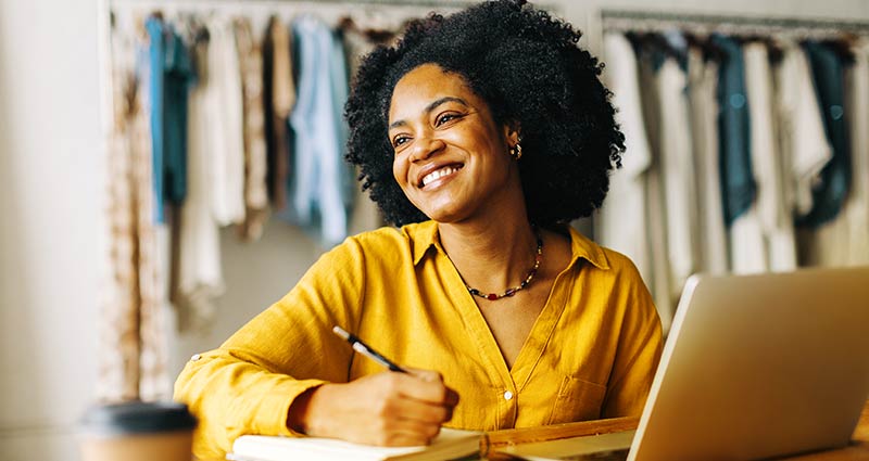 A smiling woman sits at her desk, engaged with her laptop, radiating positivity and focus in her workspace.