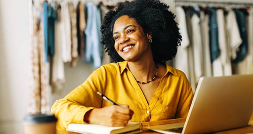 A smiling woman sits at her desk, engaged with her laptop, radiating positivity and focus in her workspace.