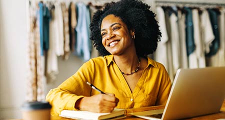 A smiling woman sits at her desk, engaged with her laptop, radiating positivity and focus in her workspace.