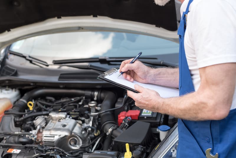 A man examines a car engine while taking notes on a clipboard, focused on his task.