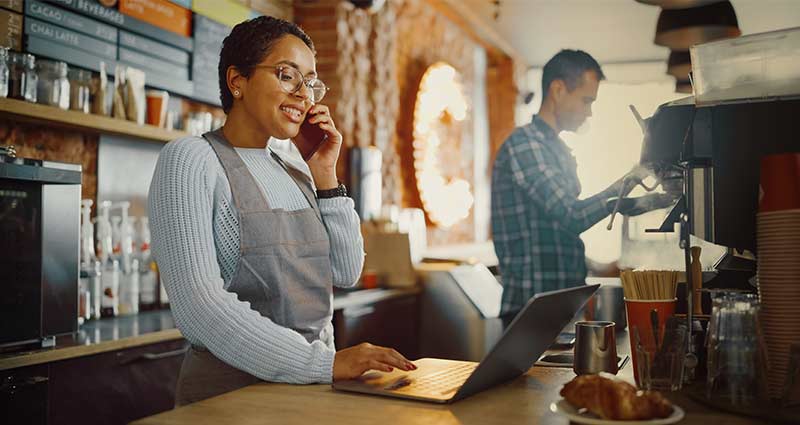 woman in cafe behind the counter working on laptop while talking on the phone, a coworker beside her is making a coffee