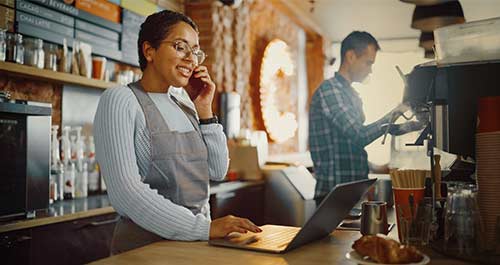 woman in cafe behind the counter working on laptop while talking on the phone, a coworker beside her is making a coffee