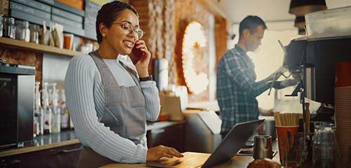 woman in cafe behind the counter working on laptop while talking on the phone, a coworker beside her is making a coffee